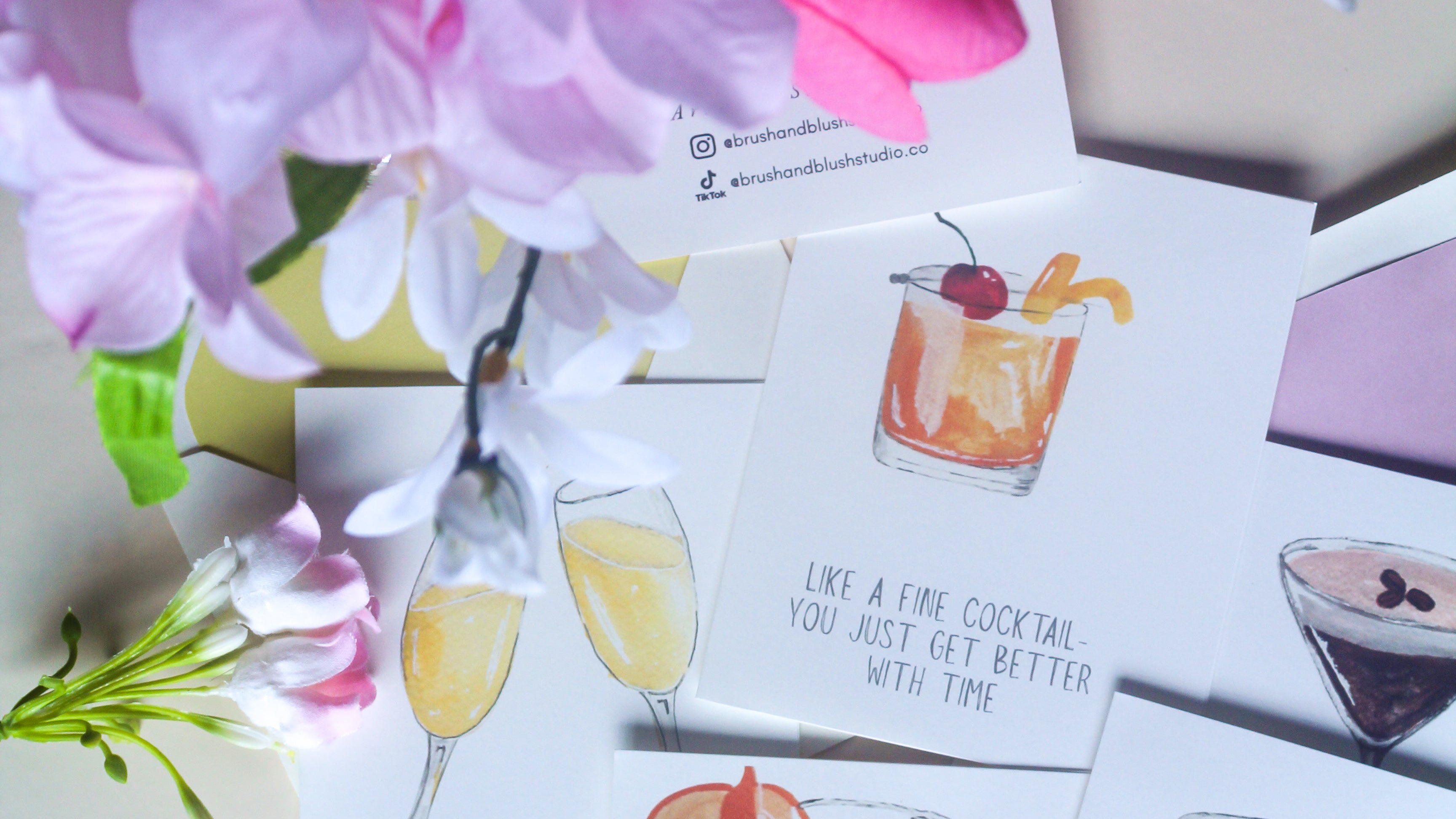 A set of illustrated cocktail-themed cards arranged on a light surface with pink and white flowers. The main card reads, 'LIKE A FINE COCKTAIL YOU JUST GET BETTER WITH TIME,' and shows a watercolor-style orange cocktail glass with a cherry and orange peel.